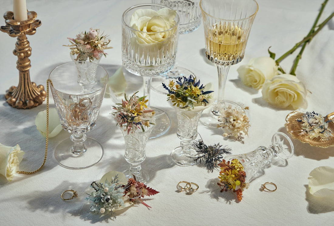 An arrangement of crystal glasses holding dried flowers and jewelry, with white roses, gold rings, a candlestick, and a pearl necklace on a white tablecloth.
