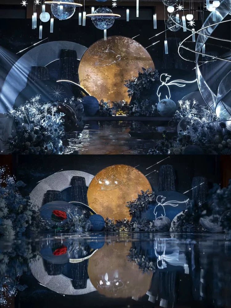 The "Little Prince Wedding Flower Rings" by CAMELLIA feature a celestial-themed setup handcrafted in New York, boasting a golden moon backdrop, abstract decor, blue lighting, and dried floral arrangements. Shimmering reflections on the glossy floor complete this enchanting stage.
