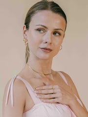 A woman with light hair poses against a neutral background, adorned in CAMELLIA's 14K Yellow Gold Vermeil Lady Amethyst Ring, wearing a pink sleeveless top, her hand gracefully resting on her chest.