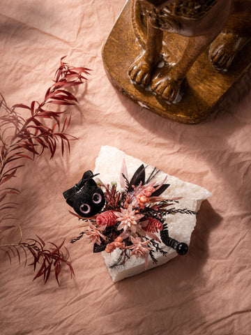 The CAMELLIA Cat Flower Ring, a small black figurine adorned with pink and red flowers, is placed on a white textured surface. To the left are dried flowers on a leafy branch, and in the upper right corner sits a carved wooden pedestal. The CAMELLIA Cat Flower Ring, a small black figurine adorned with pink and red flowers, is placed on a white textured surface. To the left are dried flowers on a leafy branch, and in the upper right corner sits a carved wooden pedestal.