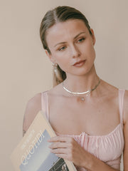 A woman with light brown hair holds a magazine titled "Italy Segreta Quotid" and wears a pink top with a sterling silver necklace. Her look is elegantly enhanced by Elsa Peony Sapphire Earrings from CAMELLIA, handcrafted in New York, against a neutral background.
