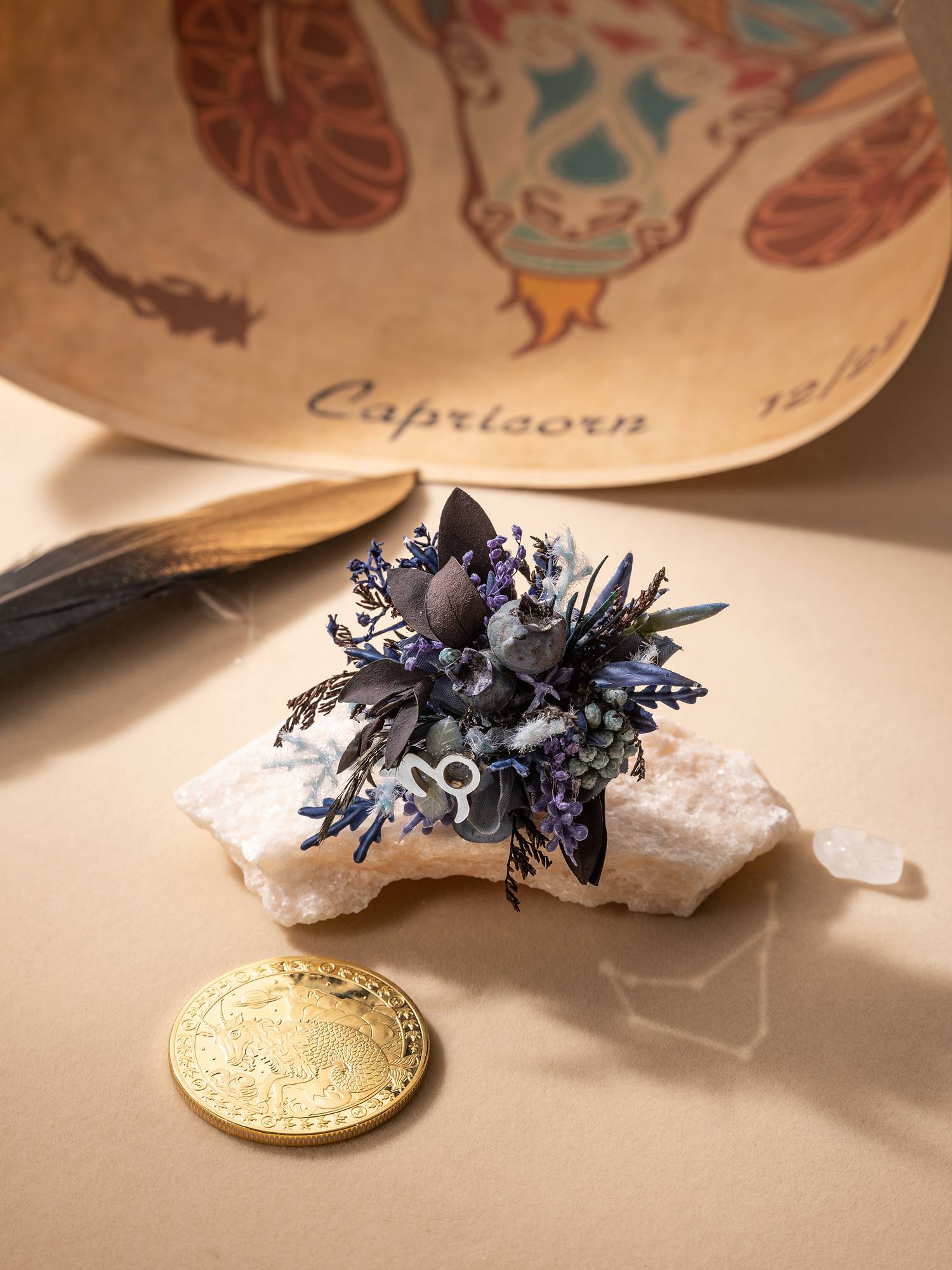 A small bouquet with dark leaves and purple flowers rests on a stone, set against a Capricorn-themed backdrop. The Capricorn Flower Ring by CAMELLIA glimmers nearby, accompanied by a feather, gold coin, and translucent pebble in this enchanting scene.