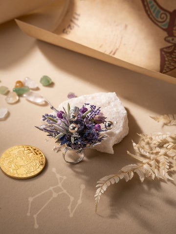 The CAMELLIA Sagittarius Flower Ring, a handcrafted decorative piece with purple, blue, and silver elements, is displayed on a white rock. The ring's adjustable size guarantees the perfect fit. Surrounding it are a gold coin, assorted crystals, and dried ferns on a beige surface. The CAMELLIA Sagittarius Flower Ring, a handcrafted decorative piece with purple, blue, and silver elements, is displayed on a white rock. The ring's adjustable size guarantees the perfect fit. Surrounding it are a gold coin, assorted crystals, and dried ferns on a beige surface.
