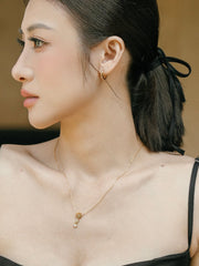 Against a neutral background, a woman with dark hair tied back in a black top and gold hoop earrings wears CAMELLIA's 15mm Mother Hoops and a 14K Yellow Gold Vermeil necklace, looking to the side.