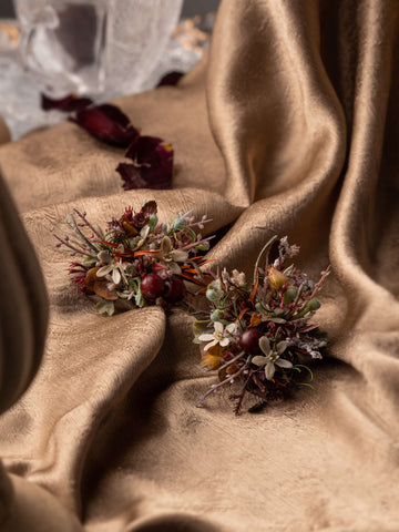 Two CAMELLIA Medieval Gothic Wedding Flower Rings featuring red berries and greenery rest on crumpled golden fabric, with a crystal glass in the background. Two CAMELLIA Medieval Gothic Wedding Flower Rings featuring red berries and greenery rest on crumpled golden fabric, with a crystal glass in the background.