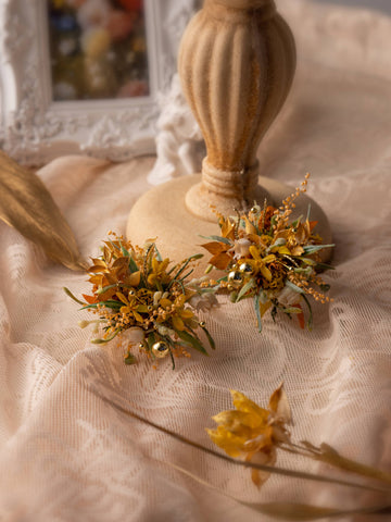 The "Love In The Sunset Wind Wedding Flower Rings" by CAMELLIA feature yellow and orange flowers, greenery, and gold accents displaying a lace tablecloth. A beige candleholder enhances the elegant scene, with dried flowers adding rustic charm beside a blurred photo in the background. The "Love In The Sunset Wind Wedding Flower Rings" by CAMELLIA feature yellow and orange flowers, greenery, and gold accents displaying a lace tablecloth. A beige candleholder enhances the elegant scene, with dried flowers adding rustic charm beside a blurred photo in the background.