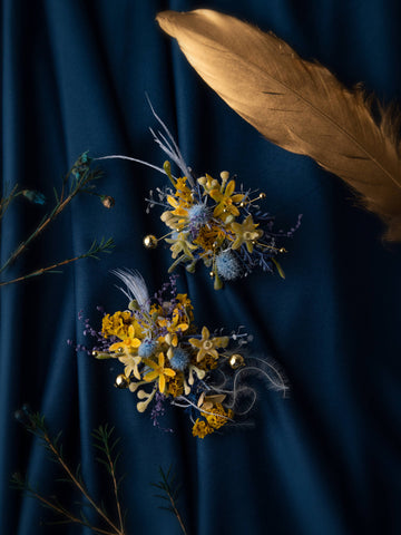 Two Van Gogh's Starry Wedding Flower Rings by CAMELLIA, featuring yellow, blue, and purple flowers on dark fabric with dried accents, are complemented by a large golden feather. Two Van Gogh's Starry Wedding Flower Rings by CAMELLIA, featuring yellow, blue, and purple flowers on dark fabric with dried accents, are complemented by a large golden feather.
