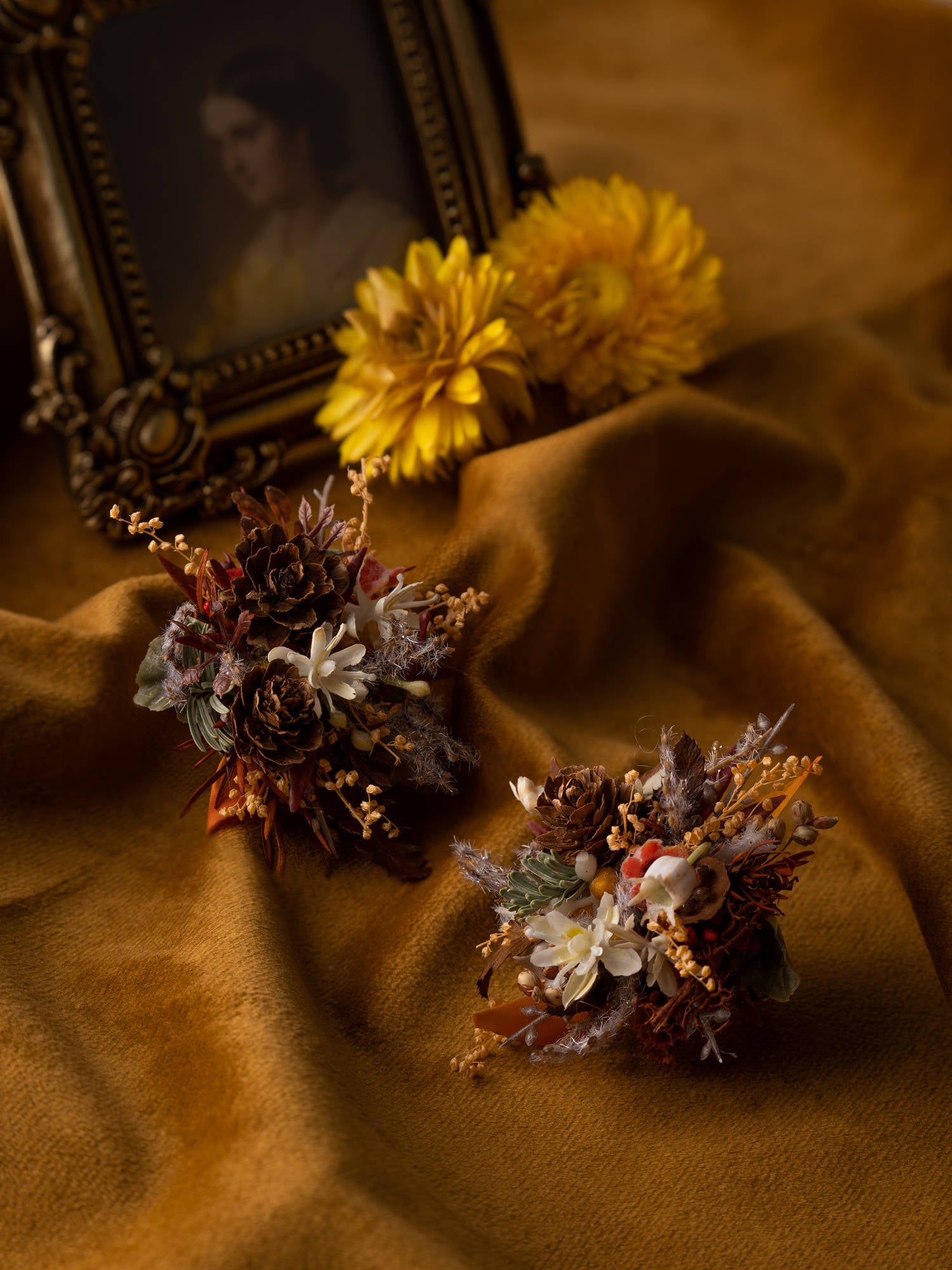 Dried flower bouquets rest on soft brown fabric beside a vintage portrait, with yellow blooms enhancing the scene. Crafted in New York, CAMELLIA's Vintage Maillard Modern Year Wedding Flower Rings add unique charm and elegance to the display with their adjustable design.