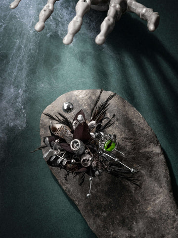 The Devil's Eye Flower Ring by CAMELLIA features an eerie decorative floral arrangement with black leaves, silver accents, and a skeleton hand on a stone slab against green. Handcrafted in New York with dried preserved flowers, it exudes an elegant charm. The Devil's Eye Flower Ring by CAMELLIA features an eerie decorative floral arrangement with black leaves, silver accents, and a skeleton hand on a stone slab against green. Handcrafted in New York with dried preserved flowers, it exudes an elegant charm.
