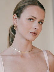 A woman with brown hair tied back, wearing CAMELLIA's Elsa Lotus Ruby 14K yellow gold vermeil earrings and a thin gold necklace, smiles slightly at the camera in a pale pink top.