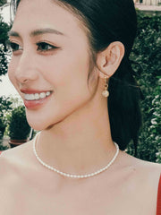 A woman outdoors, wearing CAMELLIA's Elsa Rose Pearl Earrings and gold vermeil accessories, smiles while looking to the side. Her handcrafted jewelry beautifully complements the lush plants around her.