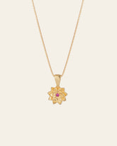 The Elsa Lotus Ruby Necklace by CAMELLIA is a handcrafted gold piece with a lotus-shaped pendant, featuring a small pink gemstone at the center, crafted in New York and displayed against a plain backdrop.