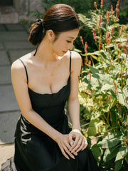 A woman in a black dress sits outdoors beside a garden, looking down with her hands clasped, her wrists adorned with the Tulip Flower Necklace by CAMELLIA glinting softly in the sunlight.