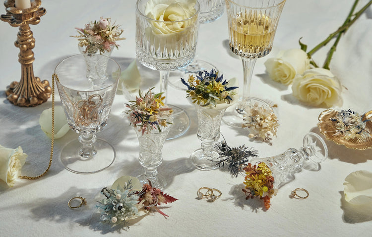 Crystal glasses, some filled with drinks and adorned with small floral arrangements, are arranged on a white tablecloth alongside white roses, gold rings, and a lit candle.