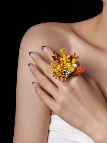 Against a dark background, a hand with painted nails showcases the Leo Flower Ring by CAMELLIA, featuring a custom symbol and adorned with yellow, orange, and brown dried preserved flowers. Against a dark background, a hand with painted nails showcases the Leo Flower Ring by CAMELLIA, featuring a custom symbol and adorned with yellow, orange, and brown dried preserved flowers.