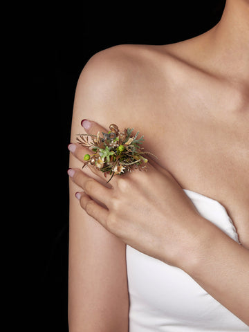 Against a black background, a person in a strapless white top holds the CAMELLIA Taurus Flower Ring—a decorative floral arrangement featuring green and gold elements, possibly with dried preserved flowers—in their left hand. Against a black background, a person in a strapless white top holds the CAMELLIA Taurus Flower Ring—a decorative floral arrangement featuring green and gold elements, possibly with dried preserved flowers—in their left hand.