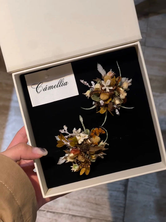A hand holds an open white box containing two small, decorative floral arrangements and a card labeled