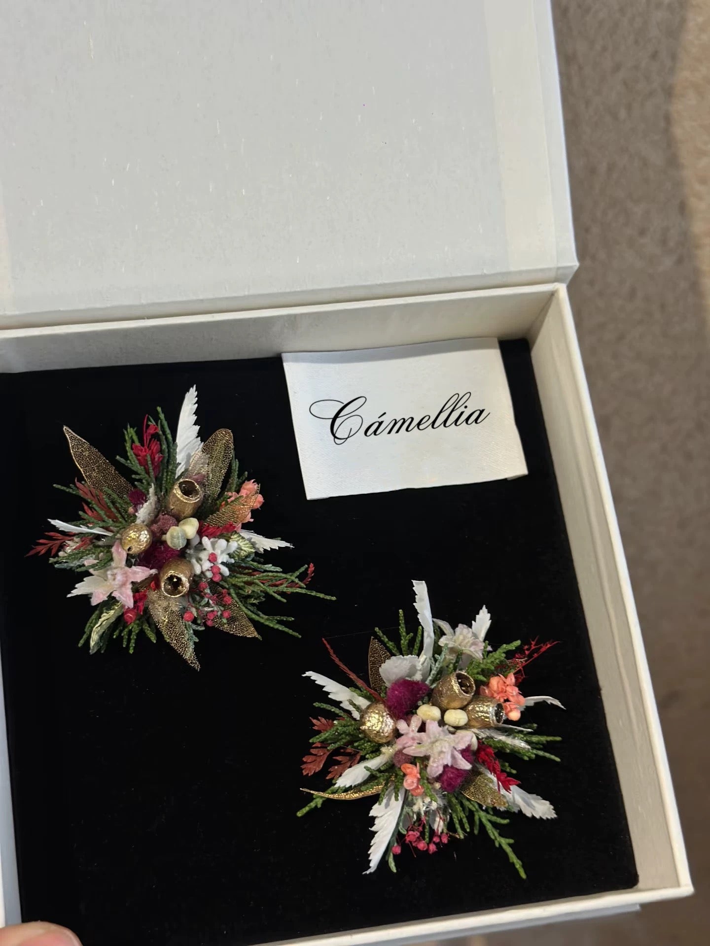 Two exquisite floral brooches lie on a black surface in an open white box, revealing the allure of handcrafted pieces. A card with "CAMELLIA" sits above them, alluding to their customizable design like that of the Customized Unique Flower Ring from CAMELLIA.