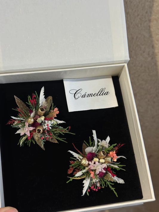 Two exquisite floral brooches lie on a black surface in an open white box, revealing the allure of handcrafted pieces. A card with