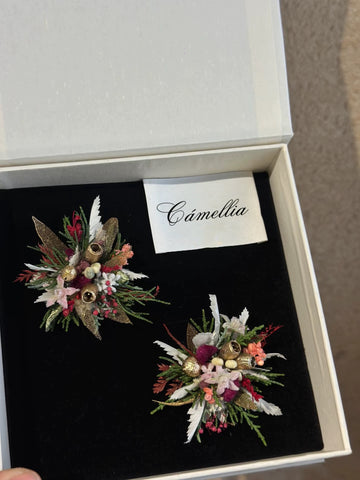 Two exquisite floral brooches lie on a black surface in an open white box, revealing the allure of handcrafted pieces. A card with "CAMELLIA" sits above them, alluding to their customizable design like that of the Customized Unique Flower Ring from CAMELLIA. Two exquisite floral brooches lie on a black surface in an open white box, revealing the allure of handcrafted pieces. A card with "CAMELLIA" sits above them, alluding to their customizable design like that of the Customized Unique Flower Ring from CAMELLIA.