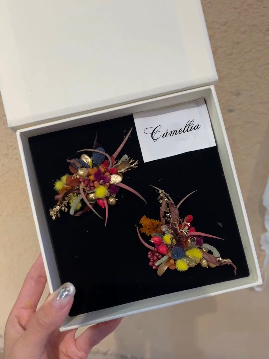 A hand holds an open box labeled "CAMELLIA," revealing two exquisite floral brooches. Each features colorful artificial flowers and leaves, showcasing the elegance of the Customized Unique Flower Ring in handcrafted jewelry.