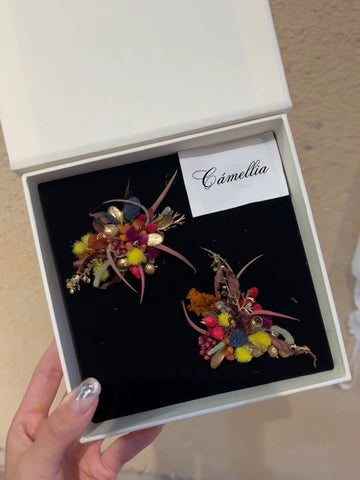 A hand holds an open box labeled "CAMELLIA," revealing two exquisite floral brooches. Each features colorful artificial flowers and leaves, showcasing the elegance of the Customized Unique Flower Ring in handcrafted jewelry. A hand holds an open box labeled "CAMELLIA," revealing two exquisite floral brooches. Each features colorful artificial flowers and leaves, showcasing the elegance of the Customized Unique Flower Ring in handcrafted jewelry.