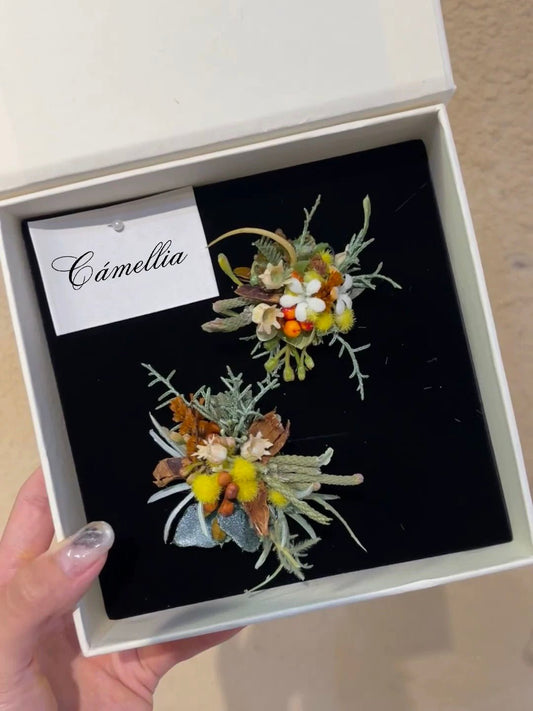 A hand holds an open box containing two intricately handcrafted floral brooches against a black background, accompanied by a card labeled