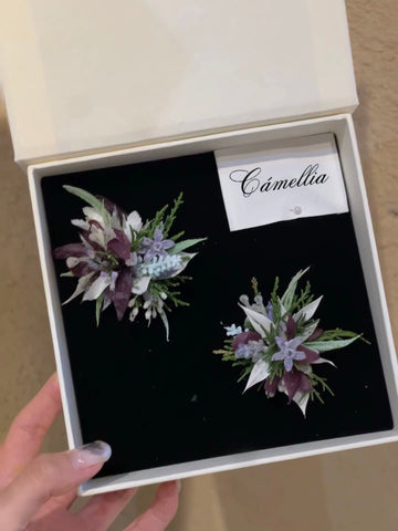A hand holds a CAMELLIA box containing two floral brooches featuring green leaves, white flowers, and purple accents. The Customized Unique Flower Ring offers a customizable design, making it perfect for any style. A hand holds a CAMELLIA box containing two floral brooches featuring green leaves, white flowers, and purple accents. The Customized Unique Flower Ring offers a customizable design, making it perfect for any style.