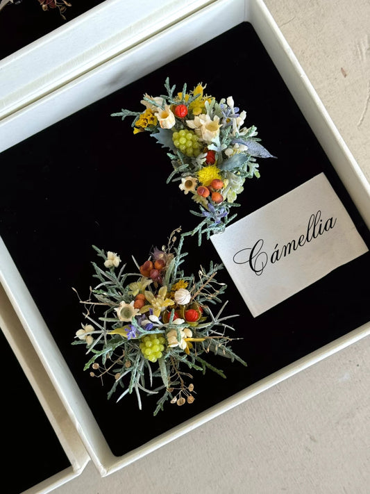 Two small, intricate floral arrangements with dried and artificial flowers displayed in a box with a card labeled