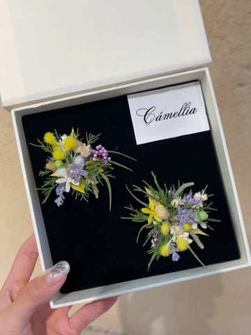 A hand holds a black box labeled "CAMELLIA," displaying two floral boutonnieres with yellow, purple, and white flowers. The customizable design of the "Customized Unique Flower Ring" echoes the elegance of handcrafted jewelry. A hand holds a black box labeled "CAMELLIA," displaying two floral boutonnieres with yellow, purple, and white flowers. The customizable design of the "Customized Unique Flower Ring" echoes the elegance of handcrafted jewelry.