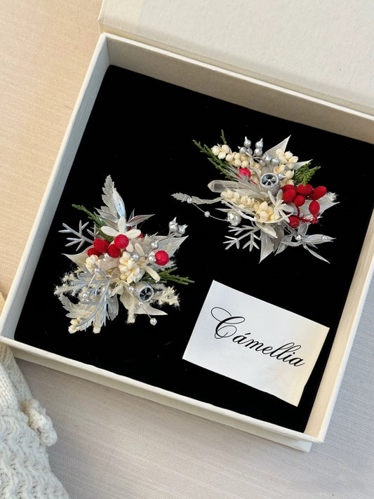 Two decorative winter-themed pins with silver leaves, red berries, and small white accents displayed in a black velvet-lined box labeled “Camellia.”. 3