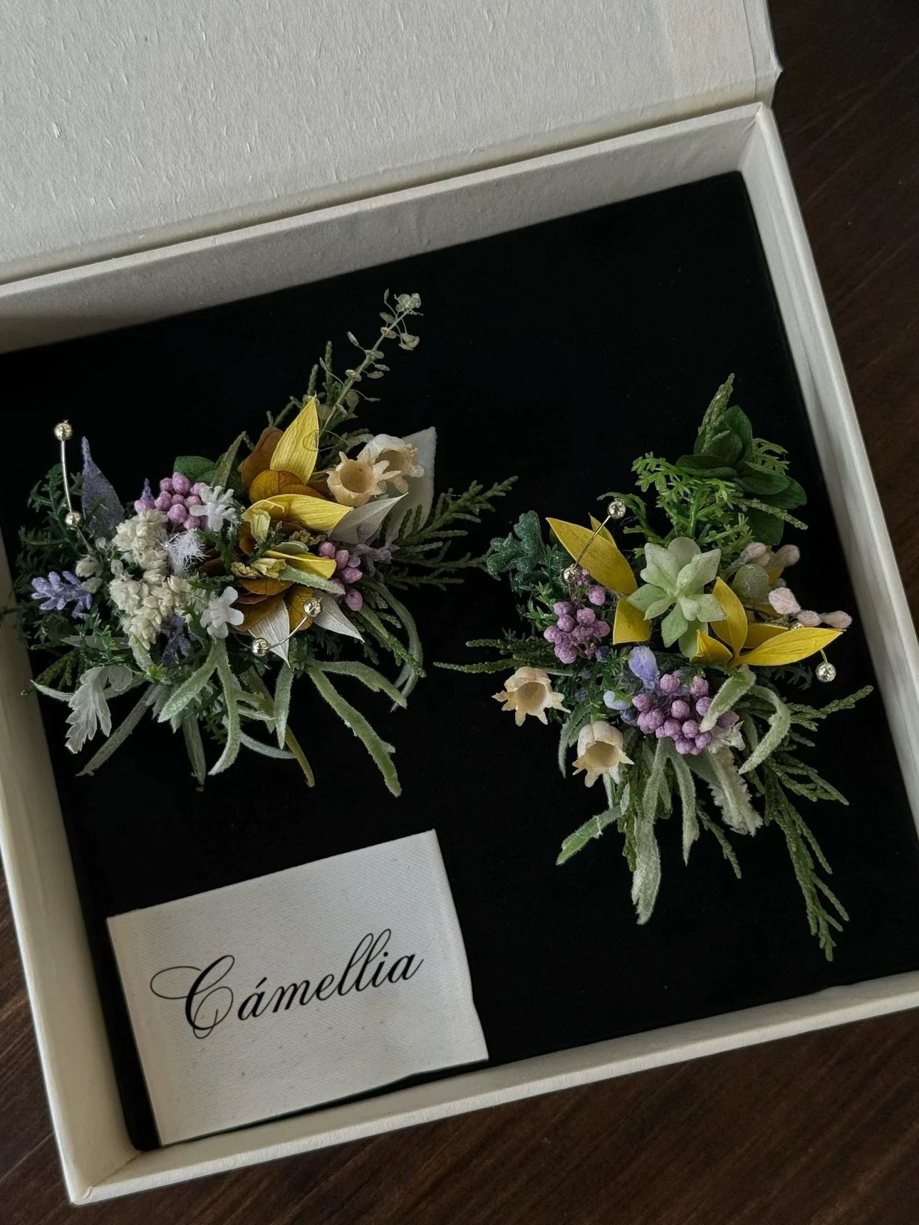 Two exquisite floral rings, featuring greenery and white, yellow, and purple flowers, are showcased on black fabric inside a white CAMELLIA box. These handcrafted jewelry pieces embody a unique charm that elevates any ensemble.
