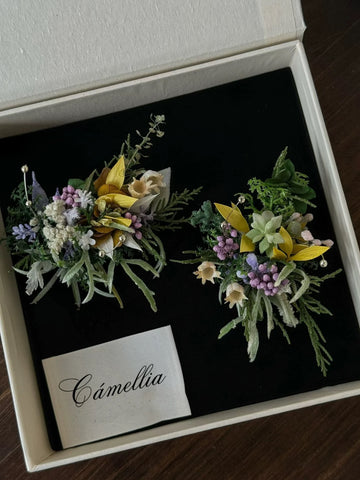 Two exquisite floral rings, featuring greenery and white, yellow, and purple flowers, are showcased on black fabric inside a white CAMELLIA box. These handcrafted jewelry pieces embody a unique charm that elevates any ensemble. Two exquisite floral rings, featuring greenery and white, yellow, and purple flowers, are showcased on black fabric inside a white CAMELLIA box. These handcrafted jewelry pieces embody a unique charm that elevates any ensemble.