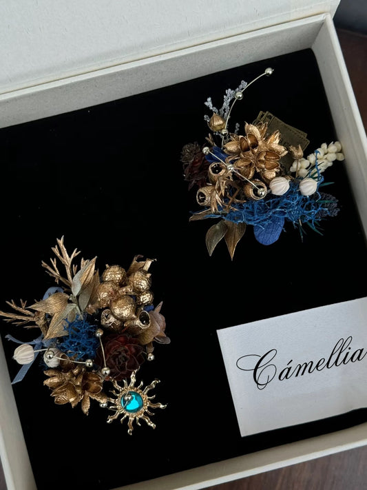 Two handcrafted jewelry brooches with gold flowers and blue accents, intricately designed, are displayed in a black velvet box labeled with the brand