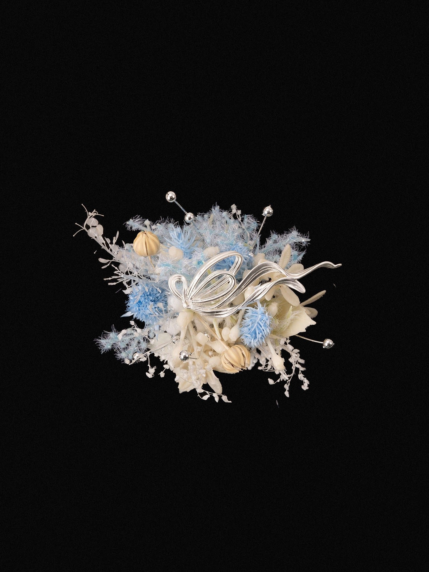 A decorative arrangement with silver metallic leaves and blue and white blooms, surrounded by delicate dried botanicals on a black background, is akin to the Ocean Flower Ring by CAMELLIA, a custom symbol flower piece handcrafted in New York.