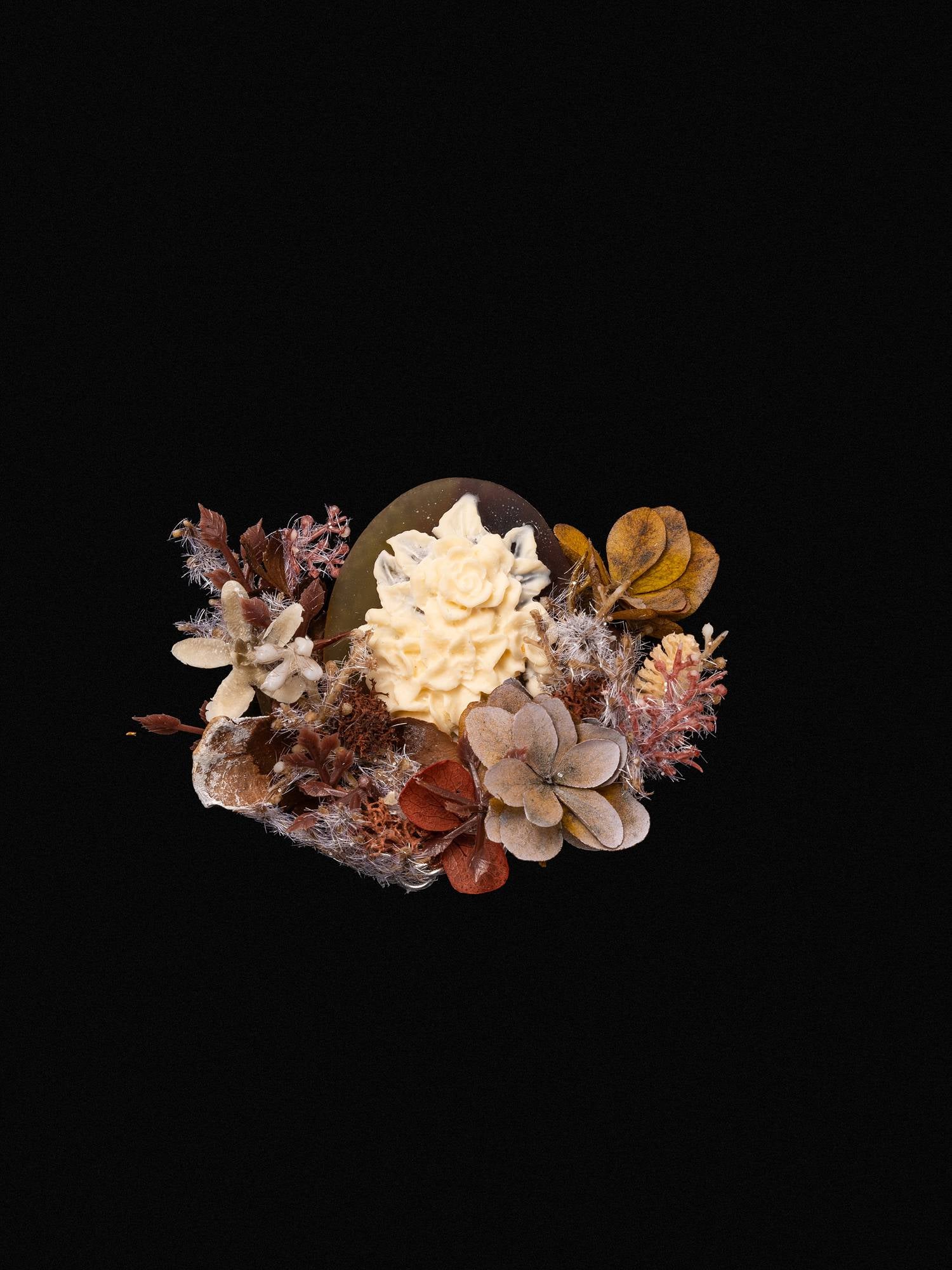 The Magnolia Birth Flower Ring by CAMELLIA, featuring an intricate ivory rose reminiscent of handcrafted jewelry, is artfully set amidst dried brown and orange leaves against a black background.