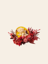 The Rose Birth Flower Ring by CAMELLIA showcases red and pink flowers encircling a floral-embossed yellow oval, set against a simple beige backdrop. Handcrafted in New York, this elegant piece adds charm to any space.