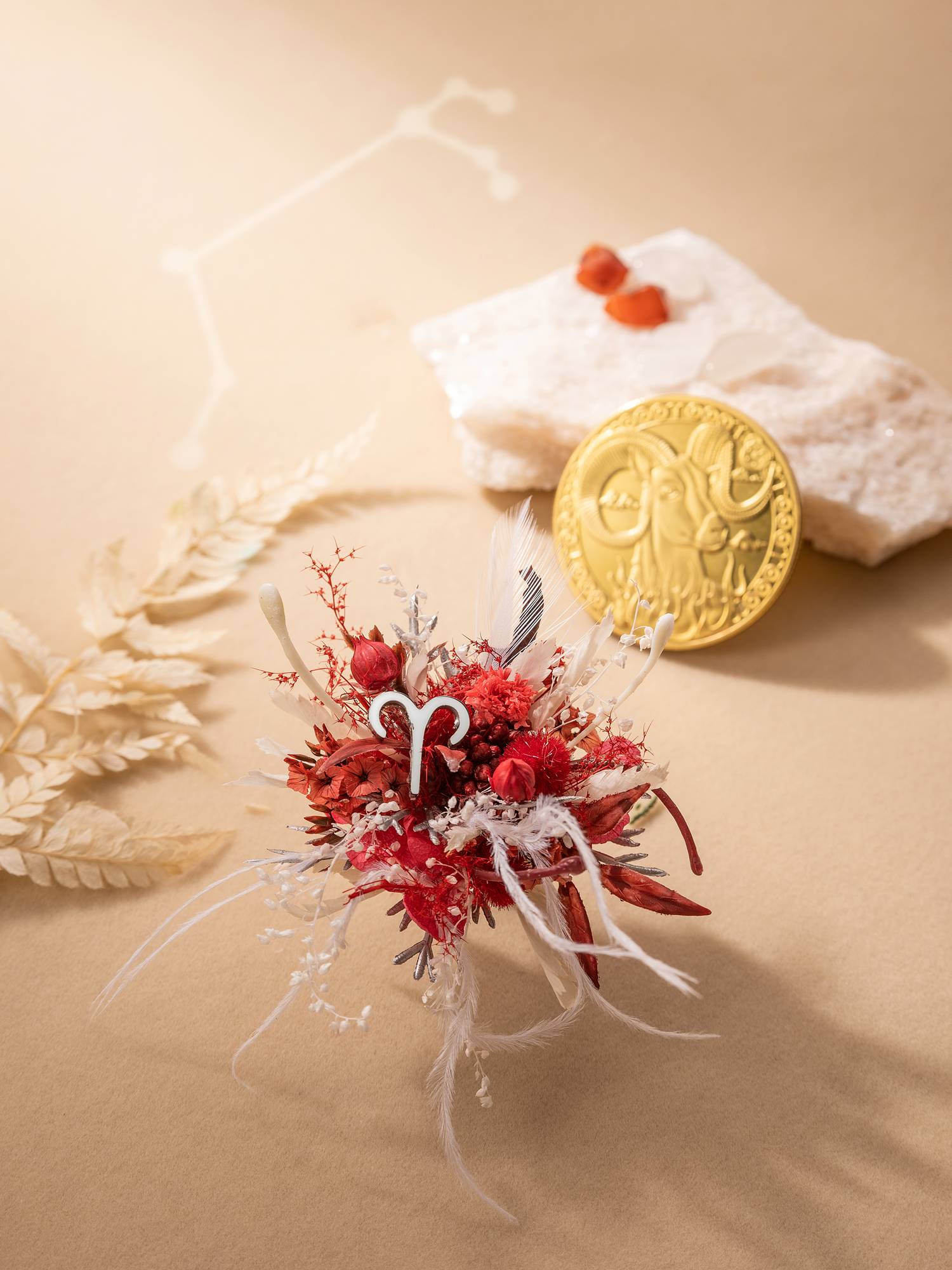 In the foreground, the Aries Flower Ring by CAMELLIA is a small handcrafted arrangement of dried red flowers and white feathers, while in the background, a gold coin, rough stones, and beige leaves rest on a light brown surface for a customizable scene with timeless appeal.