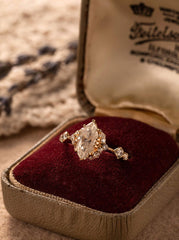 The CAMELLIA Cupid Moissanite Band, featuring a marquise-cut with surrounding moissanites, elegantly set in 14K yellow gold vermeil, is displayed in an open vintage jewelry box with luxurious red velvet lining.