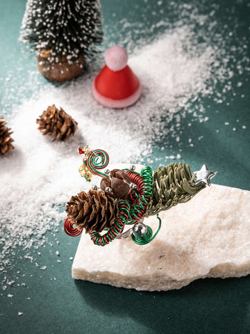 The CAMELLIA Christmas Flower Ring is a festive arrangement with pinecones, red and green spirals, and a small star on a white stone surrounded by artificial snow and miniature trees against a green background. The CAMELLIA Christmas Flower Ring is a festive arrangement with pinecones, red and green spirals, and a small star on a white stone surrounded by artificial snow and miniature trees against a green background.