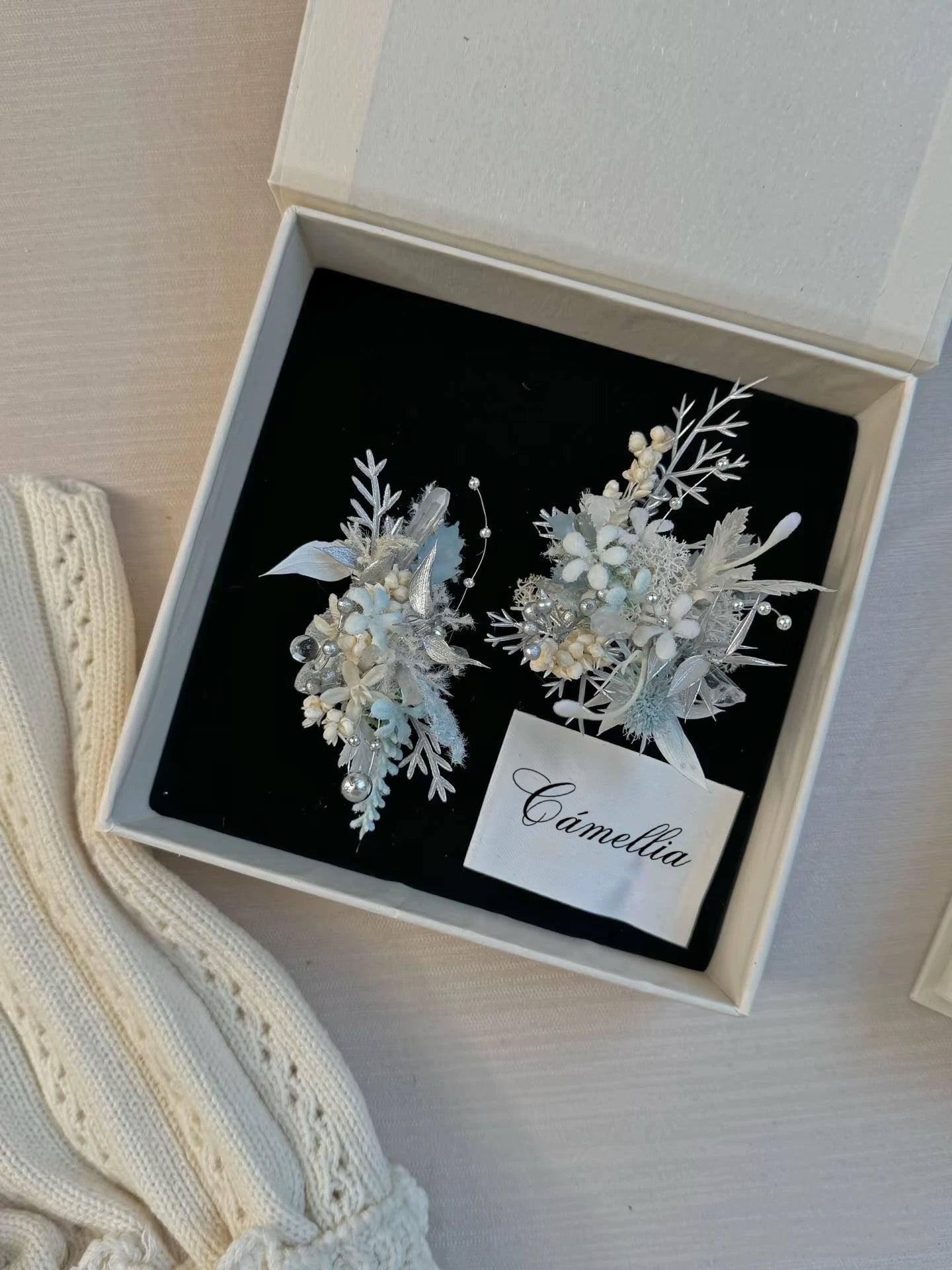 An open box revealing handcrafted jewelry with intricate white and silver floral pins on black fabric, labeled "CAMELLIA," suggests a customizable design of the Customized Holiday Flower Ring. A knitted cream fabric appears from the lower left corner.
