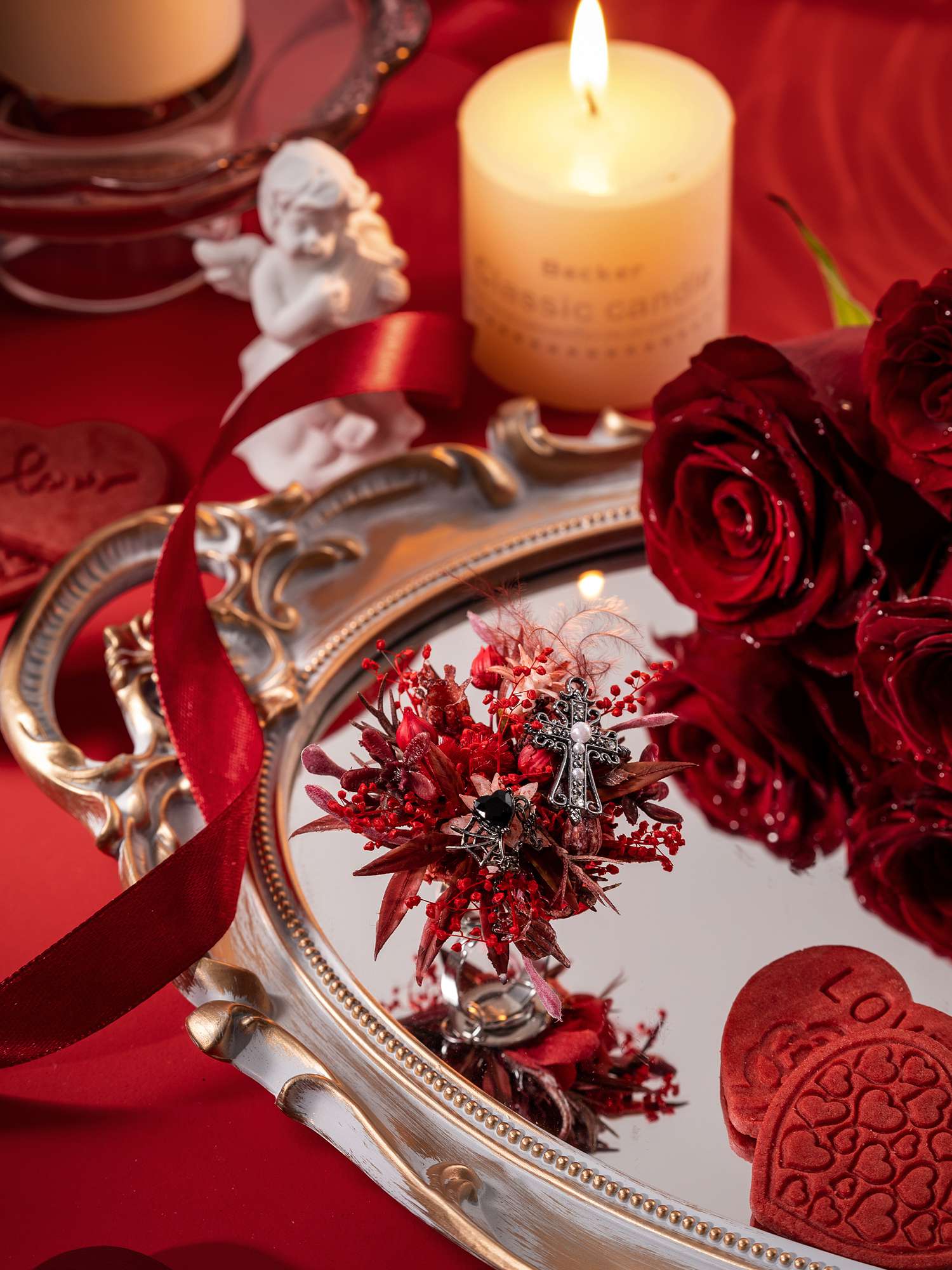 A decorative setup featuring a lit candle, angel figurine, roses, heart ornaments, and a mirrored tray reflecting red flowers and the Romantic Valentines Day Flower Ring by CAMELLIA, handcrafted in New York, all arranged on a red surface.