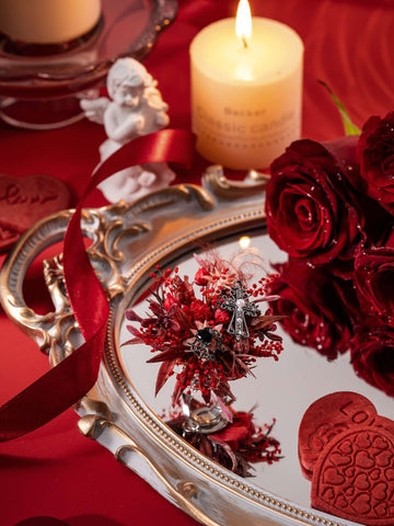A decorative setup featuring a lit candle, angel figurine, roses, heart ornaments, and a mirrored tray reflecting red flowers and the Romantic Valentines Day Flower Ring by CAMELLIA, handcrafted in New York, all arranged on a red surface. A decorative setup featuring a lit candle, angel figurine, roses, heart ornaments, and a mirrored tray reflecting red flowers and the Romantic Valentines Day Flower Ring by CAMELLIA, handcrafted in New York, all arranged on a red surface.