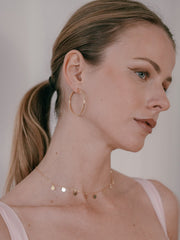 A woman with long blond hair in a ponytail wears CAMELLIA's 40mm Mother Hoops and a delicate, nickel-free hypoallergenic necklace adorned with small round pendants. She gazes to the side, exuding effortless elegance.