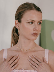 A woman with long hair in a ponytail, wearing CAMELLIA's 40mm Mother Hoops and a necklace, is dressed in a pink outfit. Her hands are near her chest as she gazes to the side.
