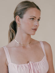 A woman with blonde hair in a ponytail wears a pink dress, CAMELLIA's 25mm Mother Hoops, and a 14K Yellow Gold Vermeil necklace, looking to the side against a neutral background.