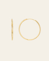 Displayed against a plain off-white background, the 40mm Mother Hoops by CAMELLIA are two gold hoop earrings made from 14K Yellow Gold Vermeil and hypoallergenic nickel-free materials. One earring highlights an elegant side view, while the other showcases its full circular shape.
