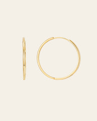 Displayed against a plain off-white background, the 40mm Mother Hoops by CAMELLIA are two gold hoop earrings made from 14K Yellow Gold Vermeil and hypoallergenic nickel-free materials. One earring highlights an elegant side view, while the other showcases its full circular shape.