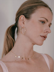 A woman with light brown hair in a ponytail wears hypoallergenic hoop earrings and CAMELLIA's Mother Chain Necklace with circular charms, glancing to the side in a softly lit environment.
