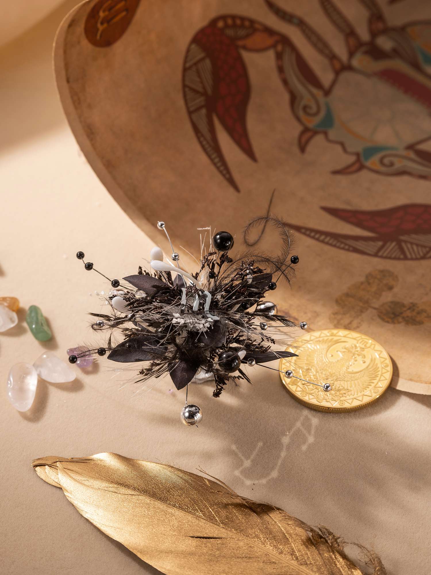 An intricate black feathered brooch with beads rests on a beige surface surrounded by colorful stones, a gold coin, and a painted crab dish. Nearby lies a golden feather next to the adjustable handcrafted Scorpio Flower Ring by CAMELLIA adorned with a custom symbol flower.